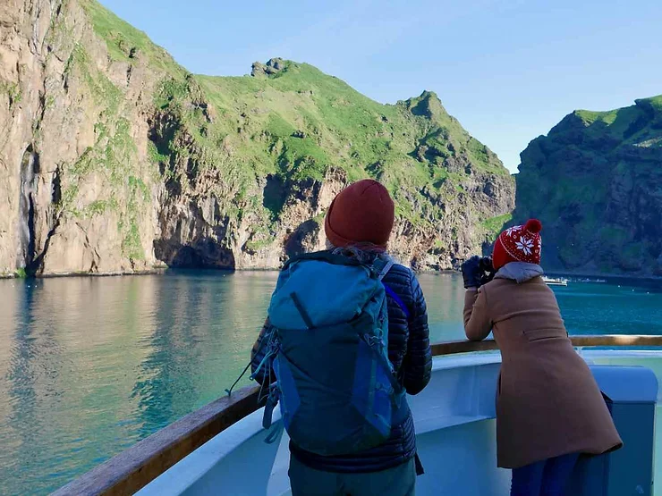 Two people on a boat tour in Iceland's Westman Islands, capturing the stunning green cliffs and calm waters with a camera on a clear day.