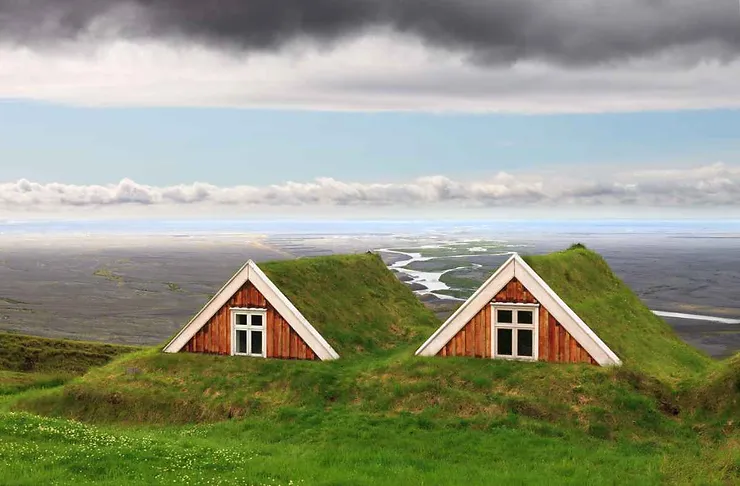 Two traditional Icelandic turf houses nestled in a grassy hillside at Skaftafell, with a river winding through the vast landscape in the background.