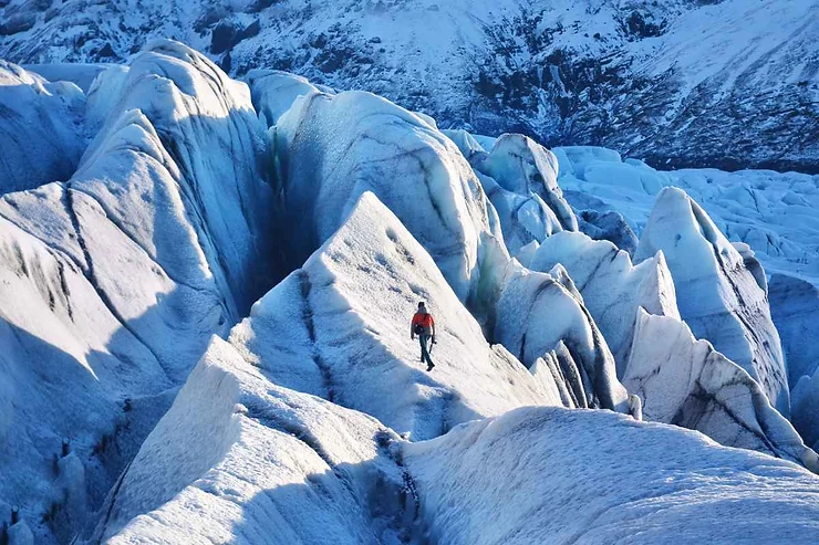 Hiker in a red jacket navigating the sharp, icy peaks of a glacier in Skaftafell, Iceland, surrounded by a vast expanse of blue ice and snow.