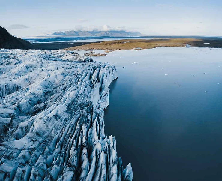 Aerial view of a glacier at Skaftafell in Iceland, with icy crevasses leading to a calm glacial lagoon and distant mountains under a clear sky.