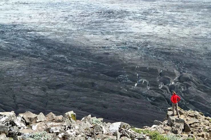 Hiker in a red jacket standing on a rocky ledge overlooking a vast glacier in Skaftafell, Iceland, showcasing the dramatic icy landscape.