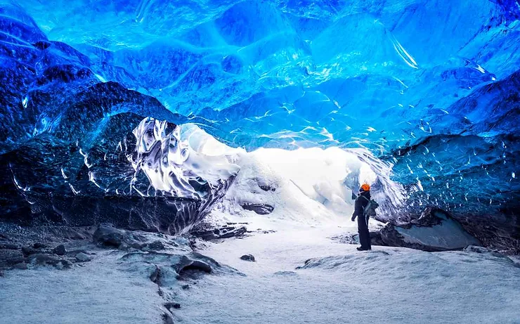 Person exploring a stunning blue ice cave in Skaftafell, Iceland, with light streaming through the entrance and illuminating the icy walls.