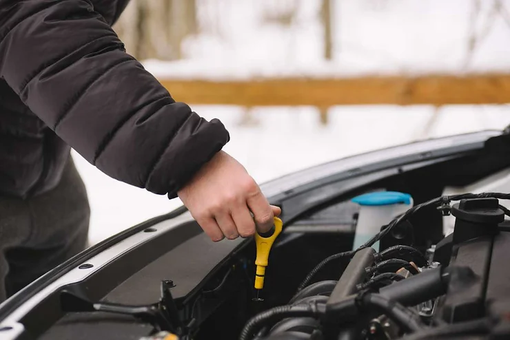 Person checking the oil level in a car engine with the hood open, preparing the vehicle for winter driving in cold weather.