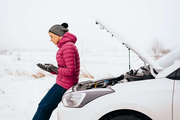 Woman in a pink jacket and winter hat standing next to a car with the hood open in snowy conditions, looking at her phone for help.