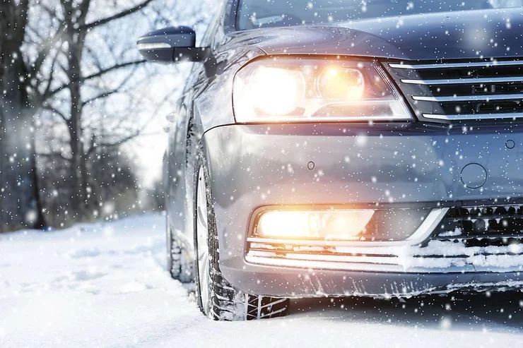 Close-up of a car driving through snow with headlights on during a snowfall, highlighting winter driving conditions.