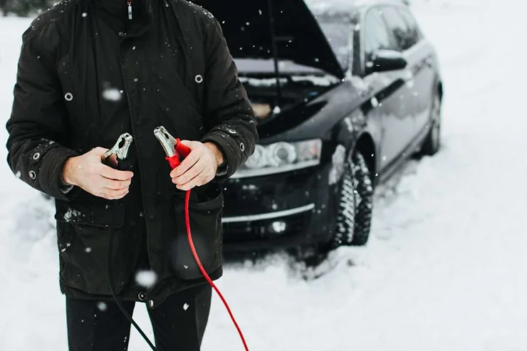 Person holding jumper cables in the snow next to a car with the hood open, preparing to jump-start a vehicle during winter.