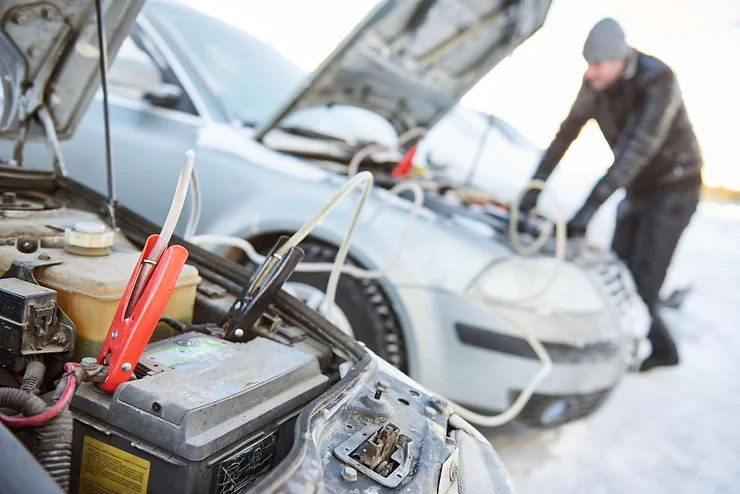 Close-up of jumper cables connected to a car battery during a winter jump-start, with a person in the background working on another vehicle.