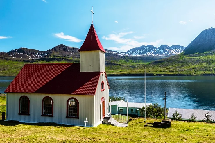 Traditional Icelandic church with a red roof in Fjardabyggd, situated by a serene lake with snow-capped mountains in the background.