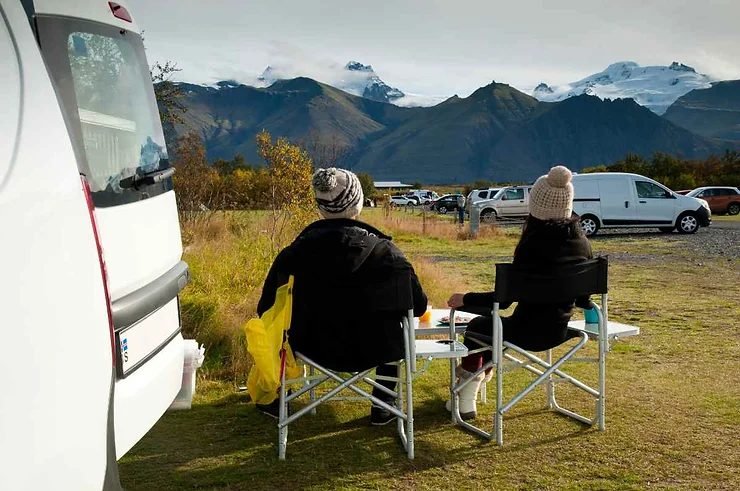 Two people sitting in camping chairs outside their vehicle, enjoying the view of mountains near Diamond Beach, Iceland.