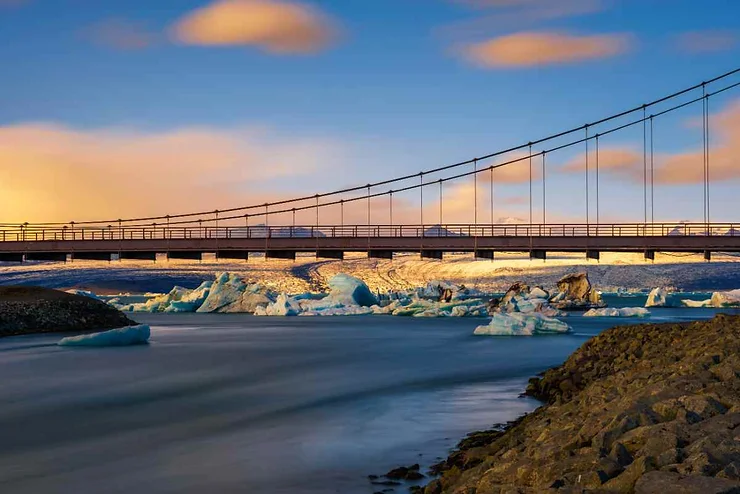 A bridge at Diamond Beach, Iceland, with icebergs floating in the water beneath during a colorful sunset.