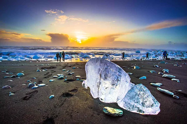 A glowing ice chunk on the black sand of Diamond Beach in Iceland during sunset, with people in the background.