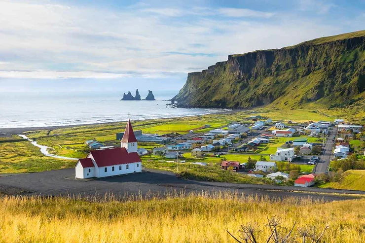 A scenic view of the town of Vík í Mýrdal, Iceland, with a church in the foreground and Reynisdrangar sea stacks in the distance.