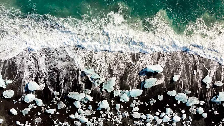 Aerial view of waves crashing onto the black sand at Diamond Beach, Iceland, with scattered icebergs on the shore.