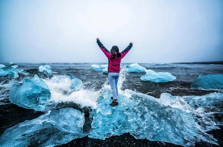 Person standing triumphantly on icebergs at Diamond Beach, Iceland, with waves crashing around them.