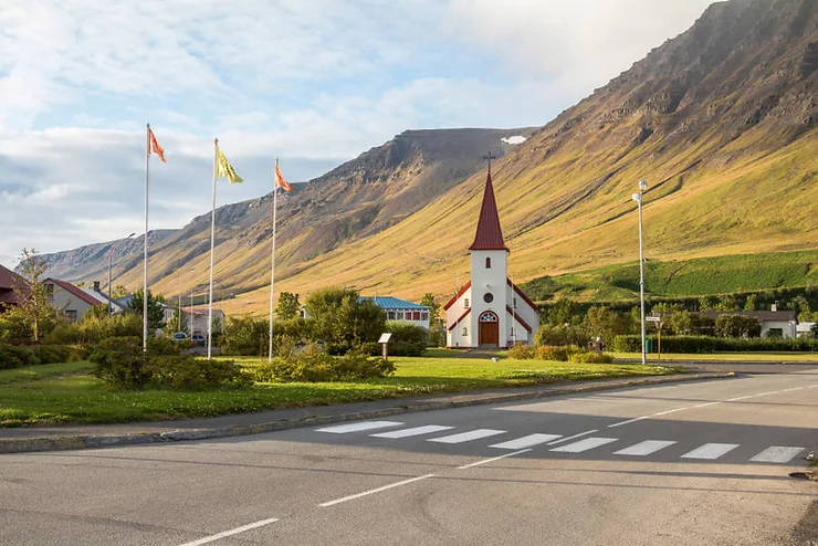 A quaint church with a red roof and steeple in the small town of Flateyri, nestled against a backdrop of towering mountains in Iceland.