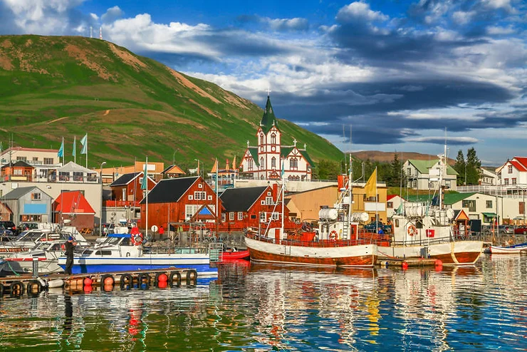 Colorful buildings and boats reflected in the water at the harbor in Husavik, Iceland, with a green hillside in the background.