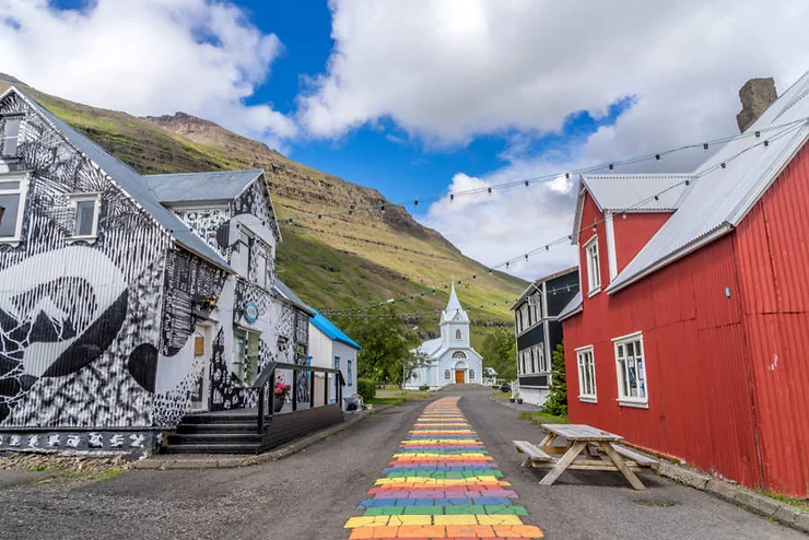 Colorful rainbow path leading to a white church in the charming town of Seydisfjordur, Iceland, with vibrant buildings on both sides and a mountainous backdrop.