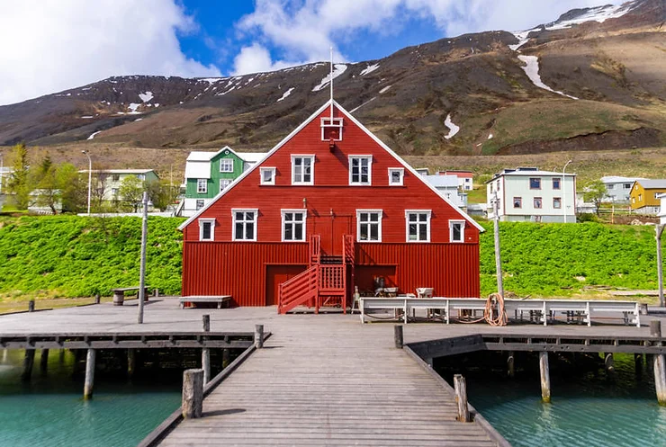 A historic red building at the Siglufjordur harbor, with colorful houses and snow-capped mountains in the background, Iceland.