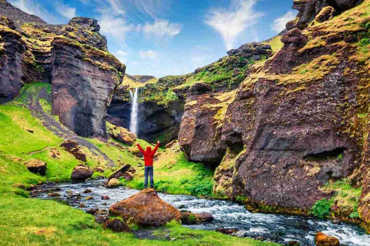 A person in a red jacket raises their arms in triumph while standing on a rock in a vibrant green valley, with a clear stream flowing towards a distant waterfall nestled between dramatic, moss-covered cliffs under a bright blue sky.