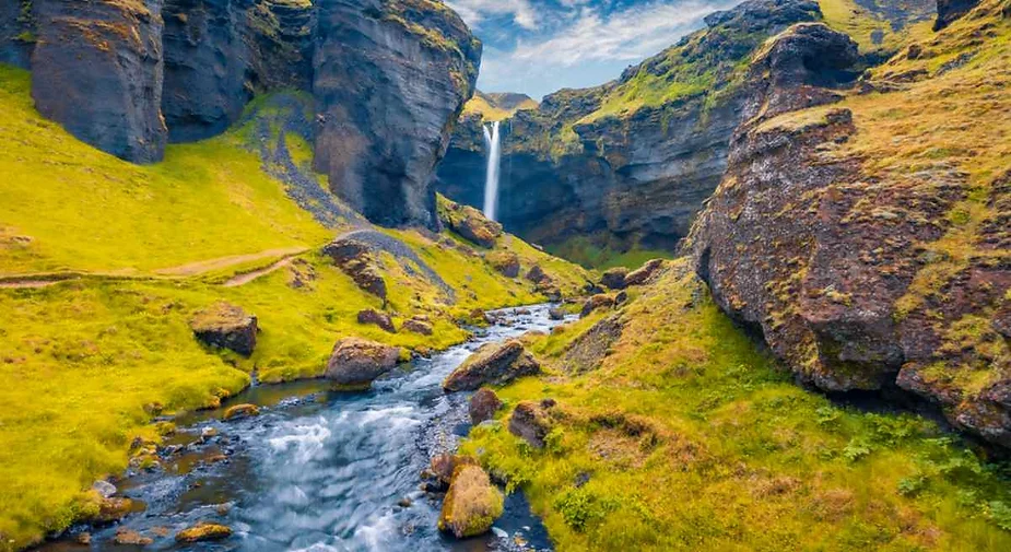 A scenic view of a peaceful river flowing through a vibrant green valley, surrounded by towering cliffs, leading to a distant waterfall cascading down into a hidden gorge under a partly cloudy sky.