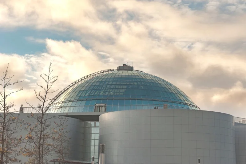 A modern, futuristic building featuring a large glass dome, reflecting the cloudy sky above, with minimalistic cylindrical structures and bare trees in the foreground.