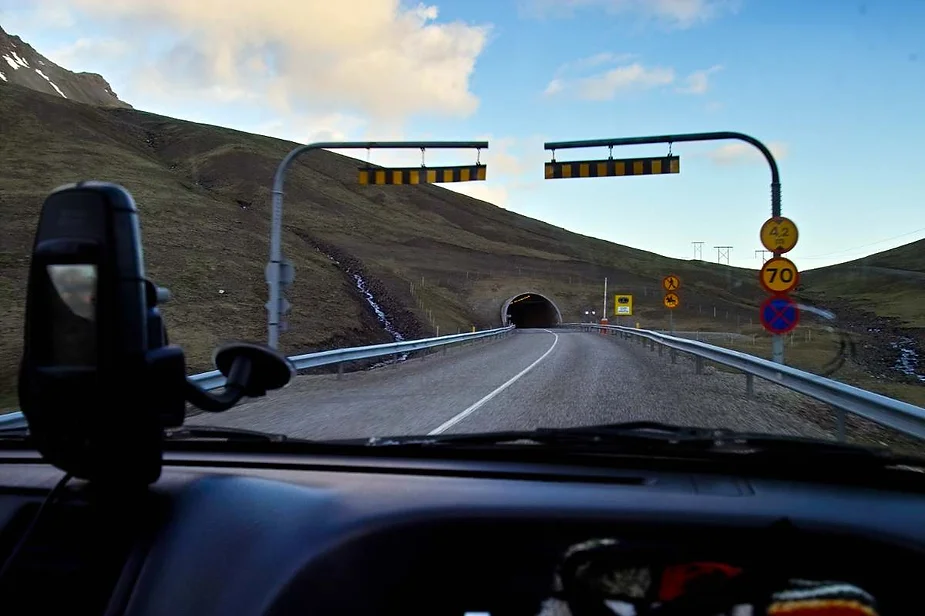 A driver's perspective from inside a car approaching a tunnel entrance on a winding rural road, with road signs and speed limits visible under a partly cloudy sky, as the vehicle moves through a mountainous landscape.