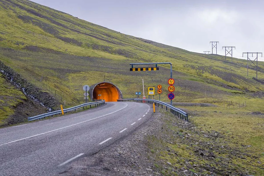 A rural road curves towards the entrance of a brightly lit tunnel cutting through a green hillside, with various road signs and speed limits posted along the way under an overcast sky.