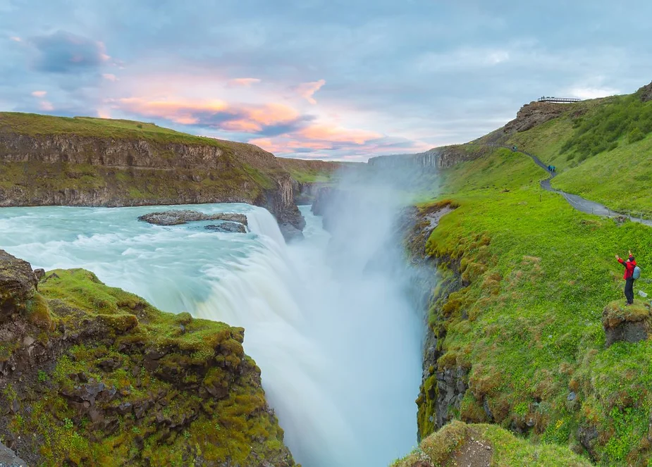 A wide-angle view of Gullfoss waterfall in Iceland, with powerful water cascading into a deep canyon surrounded by lush green cliffs, under a sky tinged with the colors of sunset. A person in red stands on the edge, capturing the scene on their phone.