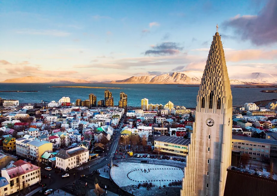 An aerial view of Reykjavik, Iceland, showcasing the iconic Hallgrímskirkja church tower in the foreground, with colorful buildings, the coastline, and distant snow-capped mountains under a clear sky.