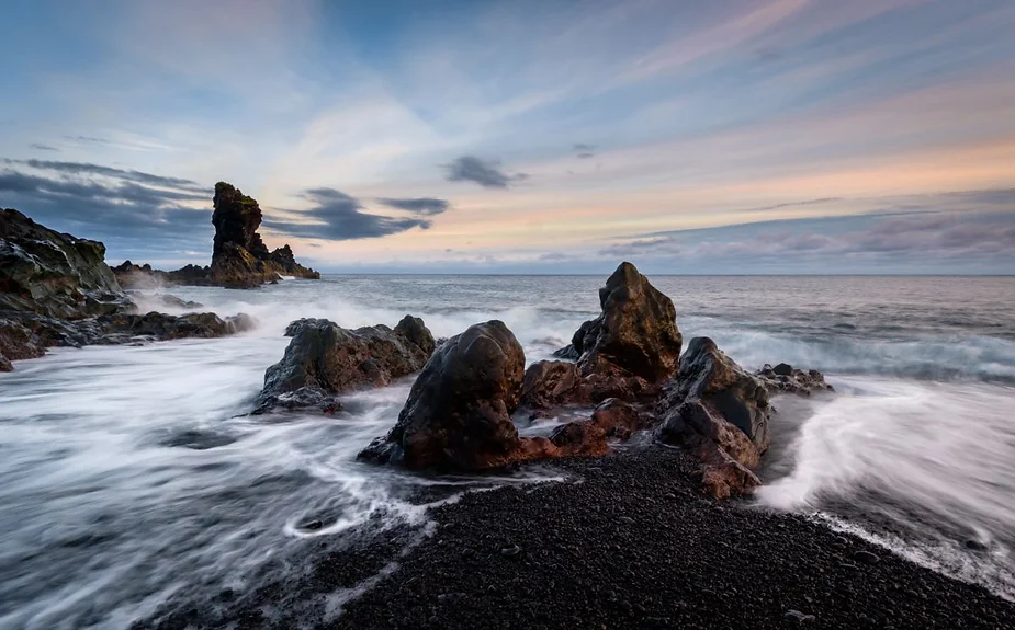 A dramatic coastal scene at Djúpalónssandur beach in Iceland, featuring rugged black volcanic rocks, waves crashing onto the dark pebbled shore, and a towering sea stack in the distance, all under a serene, pastel-colored sky at dusk.