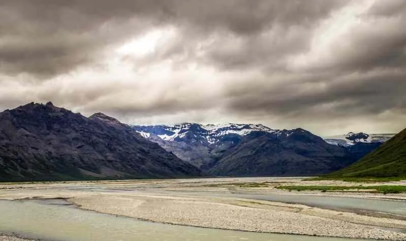 A moody landscape of a wide, braided river flowing through a valley, with rugged mountains capped with snow in the background under a cloudy, overcast sky.