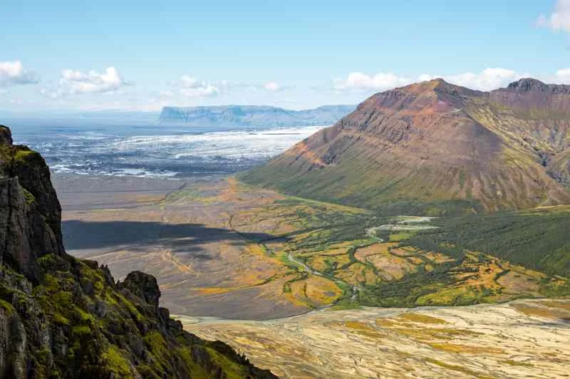 A breathtaking view of a diverse Icelandic landscape, featuring rolling hills, a glacier in the distance, and vibrant green valleys, all under a bright blue sky with scattered clouds.