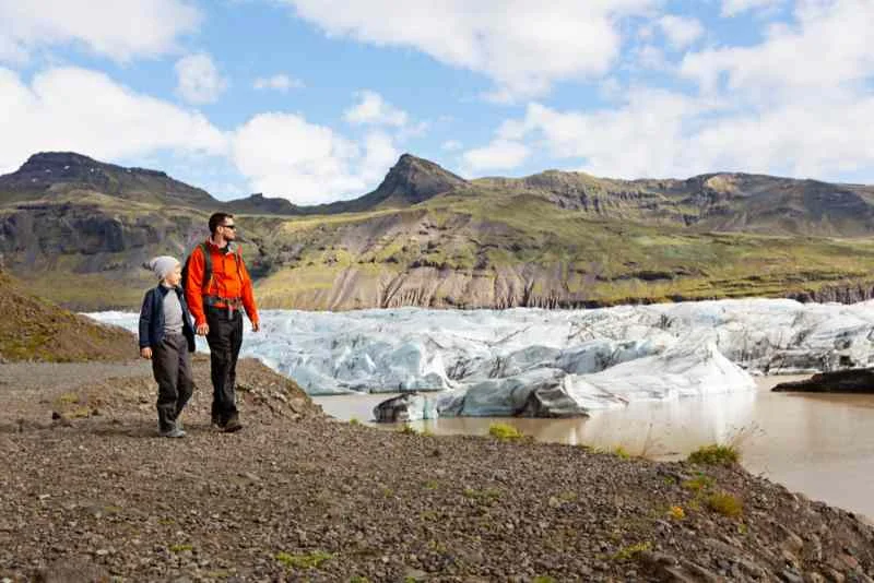 Two hikers, one wearing a red jacket and the other in a blue jacket, walk along a rocky trail beside a large glacier with towering mountains in the background, under a bright, partly cloudy sky.