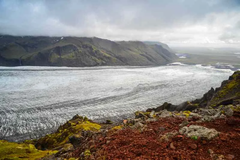 A panoramic view of a vast glacier stretching across a valley, bordered by dark, rugged mountains, with patches of vibrant green moss and red volcanic rocks in the foreground, under a cloudy sky.