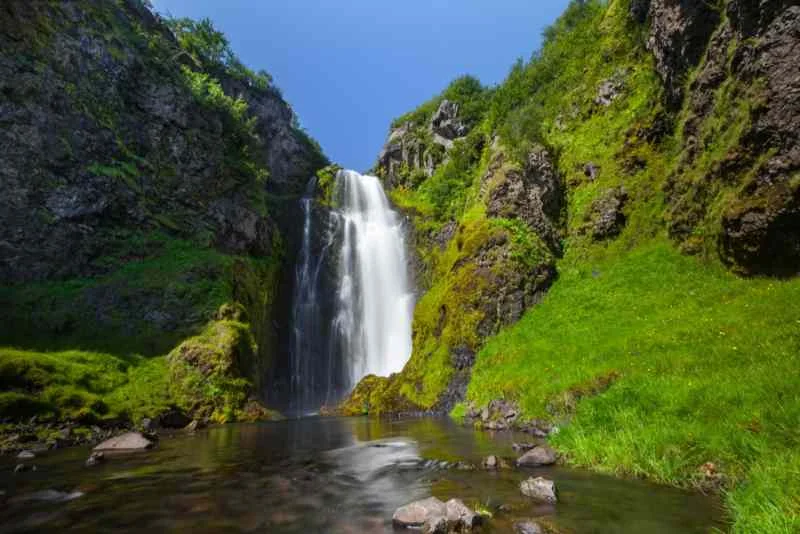 A picturesque waterfall cascades down a rocky cliff into a tranquil pool, surrounded by lush green vegetation and moss-covered rocks, under a bright blue sky.