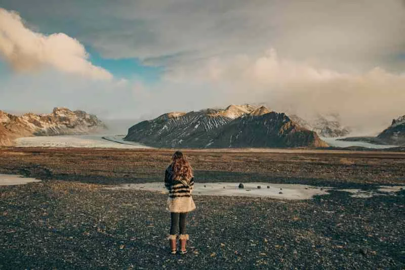 A solitary figure in a warm, striped sweater stands on a barren, rocky landscape, gazing out at a distant glacier and rugged mountain range under a dramatic, cloudy sky in Iceland.