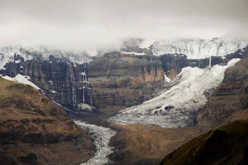 A dramatic mountain landscape featuring cascading waterfalls and glacial ice descending from steep cliffs, partially shrouded in misty clouds under a moody, overcast sky.