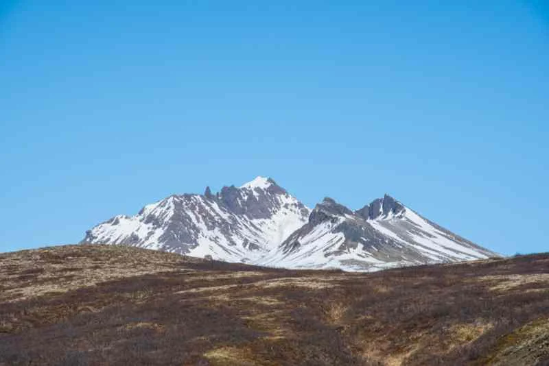 A distant view of snow-capped mountain peaks rising above a barren, brown landscape under a clear blue sky, showcasing the stark beauty of Iceland's wilderness.