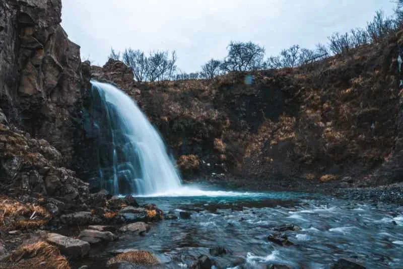 A small but powerful waterfall flows over a rocky cliff into a calm pool, surrounded by a rugged landscape with bare trees, under a cloudy sky in Iceland.