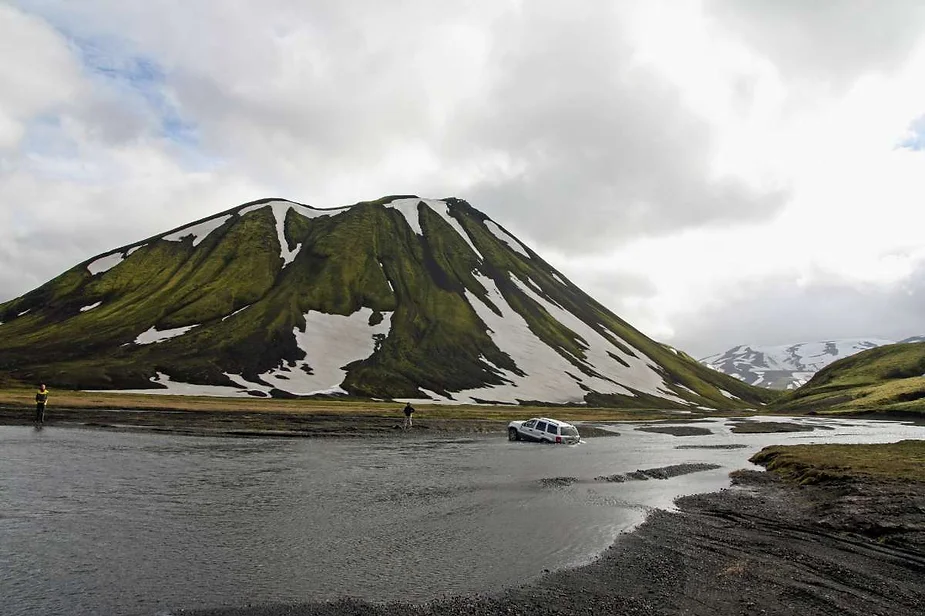 A vehicle is partially submerged in a river while attempting to cross, with a striking moss-covered mountain, accented by patches of snow, in the background under a cloudy sky in Iceland's highlands.