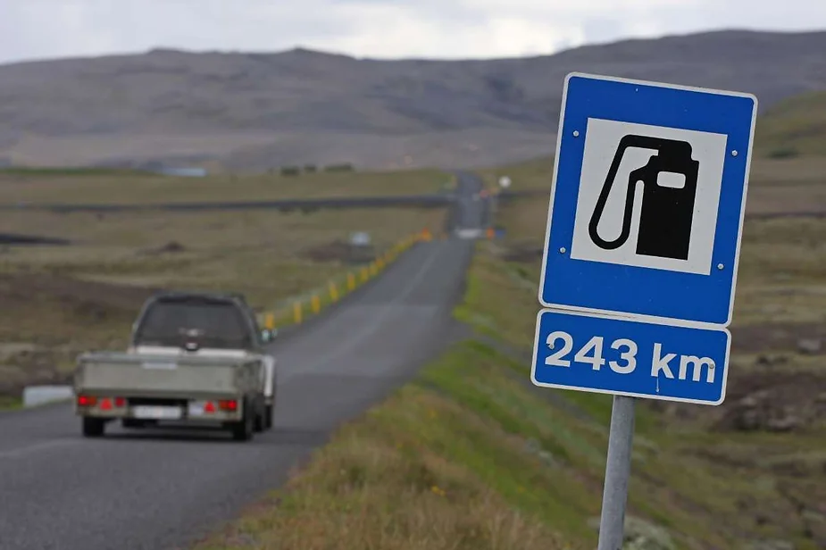 A road sign indicates that the next gas station is 243 kilometers away, as a vehicle drives down a long, empty road through a barren landscape under a cloudy sky in Iceland.