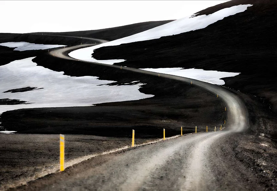 A winding gravel road snakes through a stark, barren landscape, with patches of snow clinging to the dark hillsides under a cloudy sky, creating a striking contrast between the black volcanic soil and the white snow.