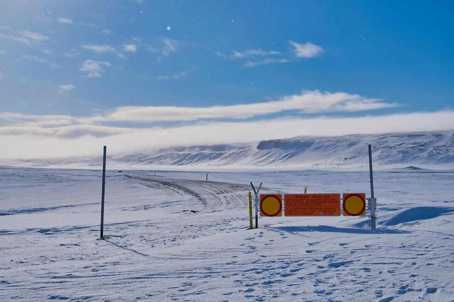 A snow-covered road in a vast, frozen landscape is blocked by a warning sign, with tire tracks leading into the distance towards snow-capped hills under a bright blue sky.