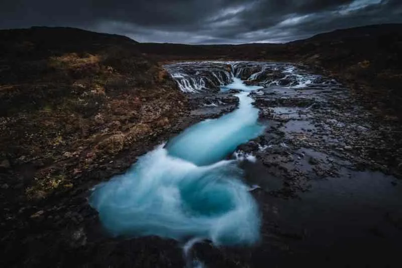 A dramatic view of Bruarfoss waterfall in Iceland under dark, moody skies, with the vibrant blue waters standing out starkly against the surrounding dark rocky landscape.