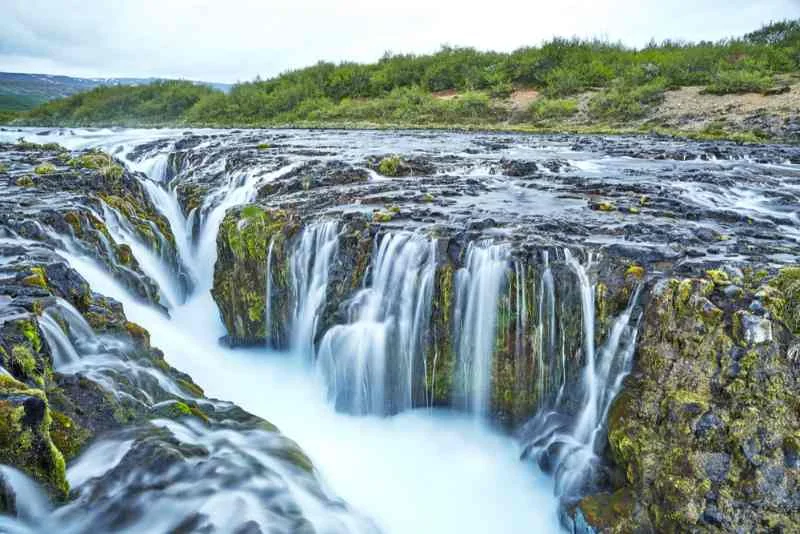 A close-up view of the cascading blue waters of Bruarfoss waterfall in Iceland, where water flows over jagged rocks into a narrow gorge, surrounded by lush greenery under a cloudy sky.