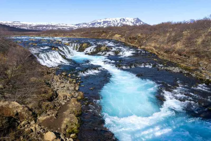 A stunning view of the vibrant blue waters of Bruarfoss waterfall in Iceland, cascading over rocks and through a narrow channel, surrounded by a rugged landscape with snow-capped mountains in the distance under a clear blue sky.