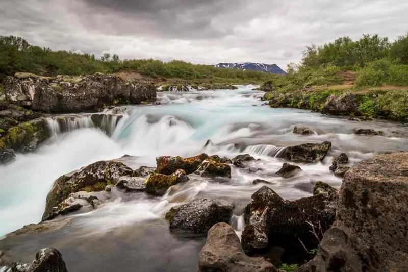 A peaceful scene of a river with turquoise water flowing over rocks and small waterfalls in a lush, green landscape under a cloudy sky in Iceland, with distant snow-capped mountains visible on the horizon.