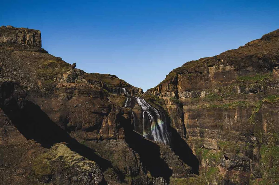 A stunning waterfall cascades down the rugged, rocky cliffs of a deep canyon in Iceland. The landscape features steep, dark brown rock faces with hints of green moss. The sky above is a clear, deep blue, creating a striking contrast with the earthy tones of the canyon.