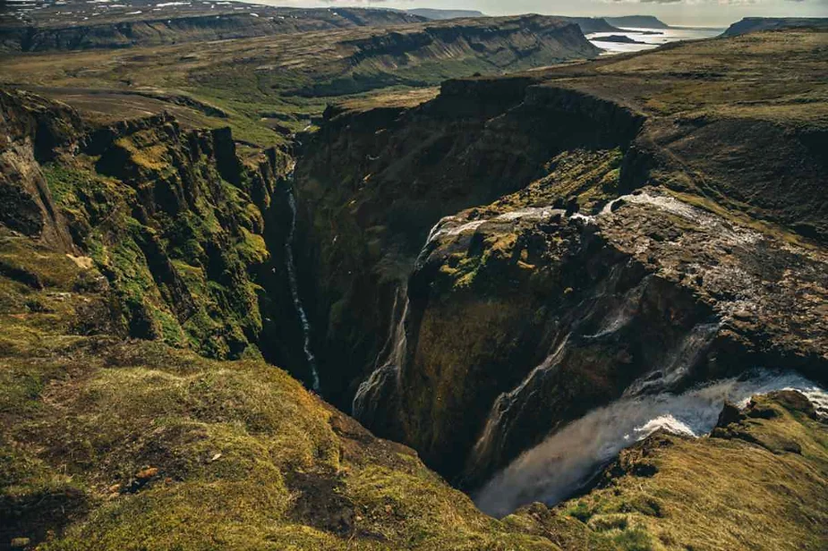 A breathtaking aerial view of a deep canyon in Iceland, where a powerful waterfall plunges into the chasm. The surrounding landscape is a mix of rugged terrain, lush green moss, and distant rolling hills under a cloudy sky.