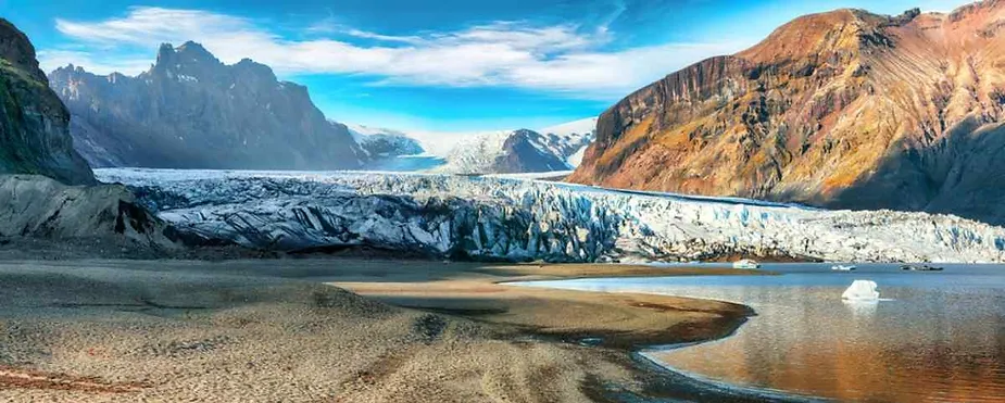 A breathtaking panoramic view of a glacier in Iceland, stretching across a rugged mountain landscape. The glacier, with its icy blue and white expanse, flows between towering, rust-colored cliffs under a vibrant blue sky with wispy clouds. In the foreground, a shallow body of water reflects the surrounding scenery, while patches of sand and rock add texture to the landscape.